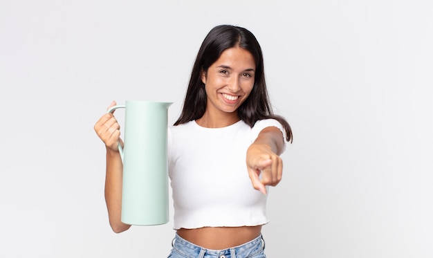 Jovem hispânica apontando para a câmera escolhendo você e segurando uma garrafa térmica de café