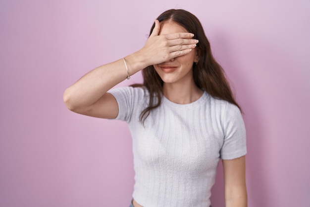 Jovem hispânica em pé sobre fundo rosa sorrindo e rindo com a mão no rosto cobrindo os olhos de surpresa. conceito cego.