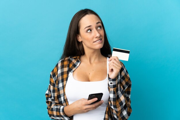 Foto jovem hispânica em uma parede azul isolada, comprando com o celular com um cartão de crédito enquanto pensa