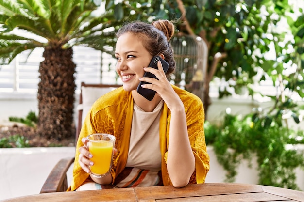 Jovem hispânica falando no smartphone e bebendo suco de laranja no terraço.