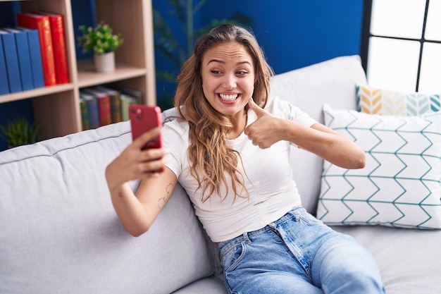 Jovem hispânica fazendo videochamada com smartphone sorrindo feliz e positivo polegar para cima fazendo sinal excelente e de aprovação