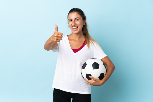 Jovem hispânica isolada azul com uma bola de futebol e com o polegar para cima