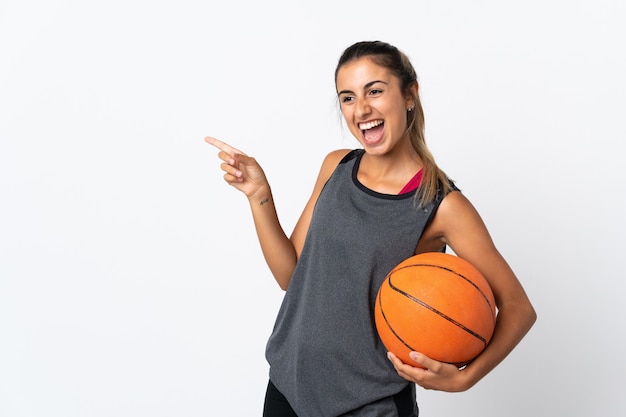 Jovem hispânica jogando basquete sobre uma parede branca isolada apontando o dedo para o lado e apresentando um produto