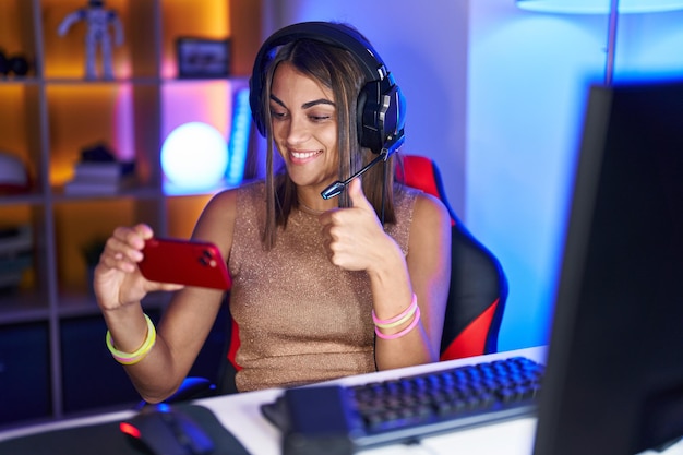 Foto jovem hispânica jogando videogame com smartphone sorrindo feliz e positivo, polegar para cima fazendo sinal excelente e de aprovação
