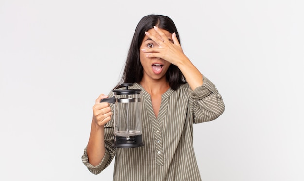 Jovem hispânica parecendo chocada, assustada ou apavorada, cobrindo o rosto com a mão e segurando uma cafeteira manual