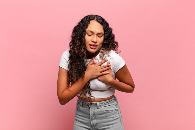 Jovem hispânica parecendo triste, magoada e com o coração partido, segurando as duas mãos perto do coração, chorando e se sentindo deprimida