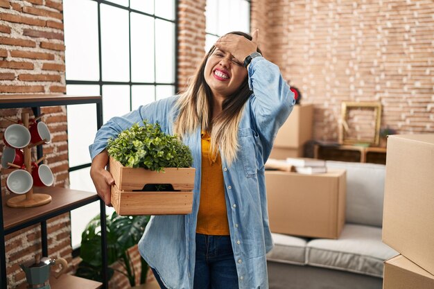 Foto jovem hispânica se mudando para uma nova casa segurando plantas estressadas e frustradas com a mão na cabeça surpresa e rosto zangado