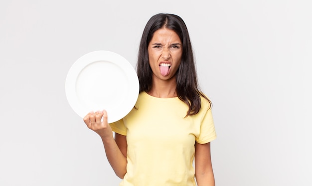 Foto jovem hispânica se sentindo enojada e irritada, com a língua de fora e segurando um prato vazio
