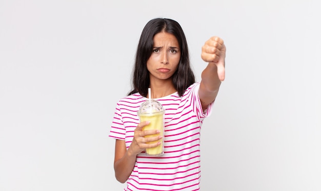 Jovem hispânica se sentindo mal, mostrando os polegares para baixo e segurando um milk-shake de baunilha