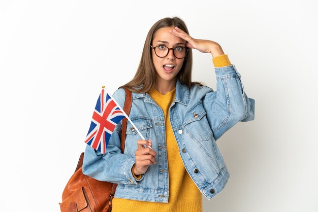 Jovem hispânica segurando uma bandeira do Reino Unido sobre um fundo branco isolado, fazendo um gesto surpresa enquanto olha para o lado