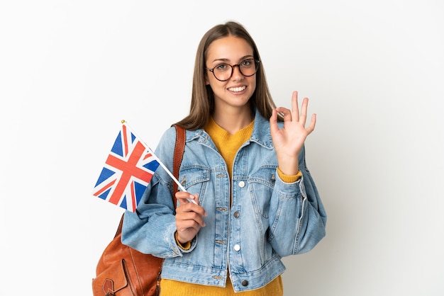 Jovem hispânica segurando uma bandeira do reino unido sobre um fundo branco isolado, mostrando um sinal de ok com os dedos