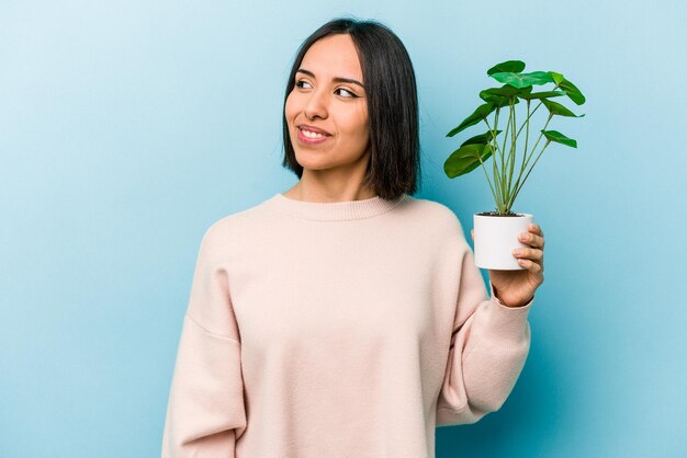 Jovem hispânica segurando uma planta isolada em fundo azul olha de lado sorrindo alegre e agradável