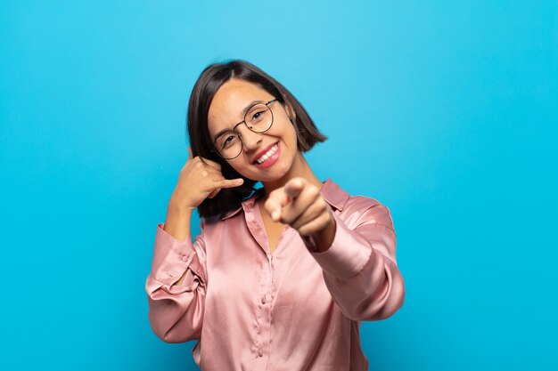 Jovem hispânica sorrindo alegremente e apontando para a frente enquanto faz um gesto de "ligar para você", falando ao telefone