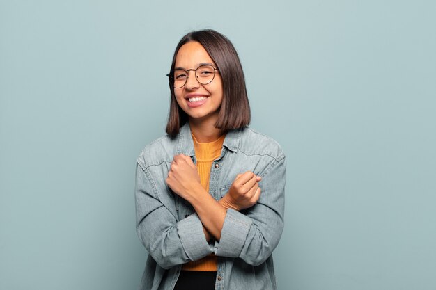 Jovem hispânica sorrindo alegremente e comemorando, com os punhos cerrados e os braços cruzados, sentindo-se feliz e positiva