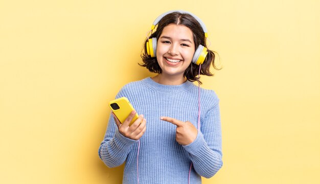 Jovem hispânica sorrindo alegremente, sentindo-se feliz e apontando para o lado. conceito de fones de ouvido e smartphone