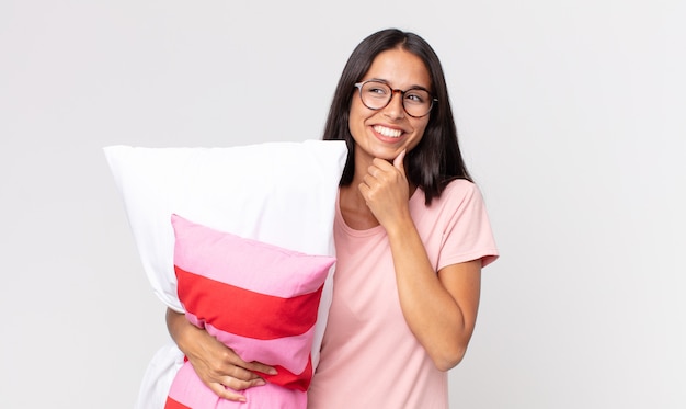 Jovem hispânica sorrindo com uma expressão feliz e confiante, com a mão no queixo de pijama e segurando um travesseiro