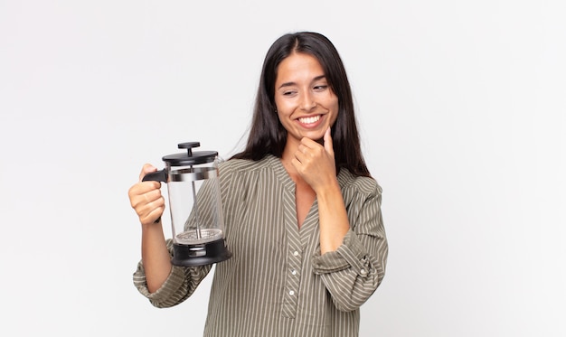 Jovem hispânica sorrindo com uma expressão feliz e confiante com a mão no queixo e segurando uma cafeteira manual