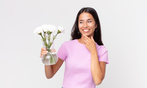 Jovem hispânica sorrindo com uma expressão feliz e confiante com a mão no queixo segurando flores decorativas