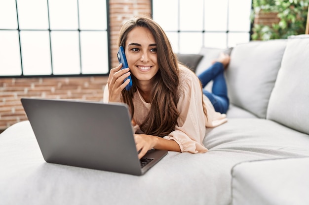 Jovem hispânica sorrindo confiante usando laptop e falando no smartphone em casa