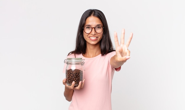 Jovem hispânica sorrindo e parecendo amigável, mostrando o número três e segurando uma garrafa de grãos de café