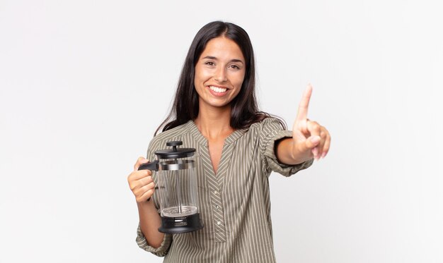 Jovem hispânica sorrindo e parecendo amigável, mostrando o número um e segurando uma cafeteira manual