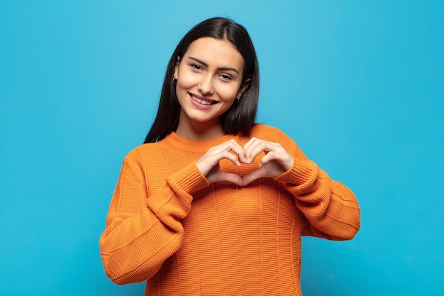 Jovem hispânica sorrindo e se sentindo feliz, fofa, romântica e apaixonada, fazendo formato de coração com as duas mãos