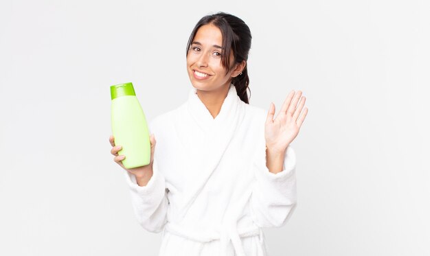 Jovem hispânica sorrindo feliz, acenando com a mão, dando as boas-vindas e cumprimentando você de roupão e segurando um shampoo