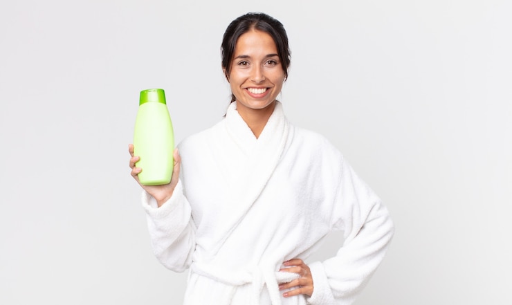Foto jovem hispânica sorrindo feliz com uma mão no quadril e confiante vestindo um roupão de banho e segurando um shampoo