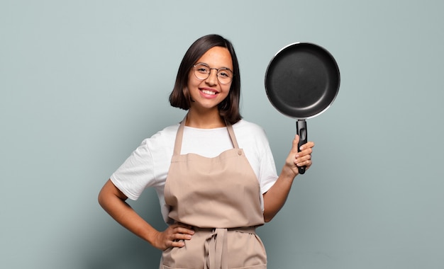 Jovem hispânica sorrindo feliz com uma mão no quadril e uma atitude confiante, positiva, orgulhosa e amigável