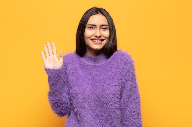 Jovem hispânica sorrindo feliz e alegre, acenando com a mão, dando as boas-vindas e cumprimentando você ou dizendo adeus