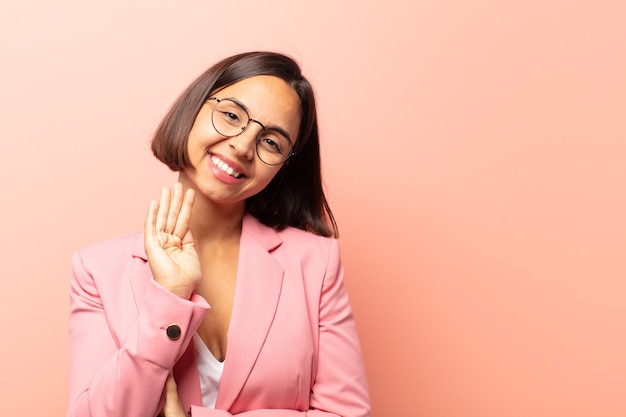 Jovem hispânica sorrindo feliz e animada, acenando com a mão, dando as boas-vindas e cumprimentando você ou dizendo adeus