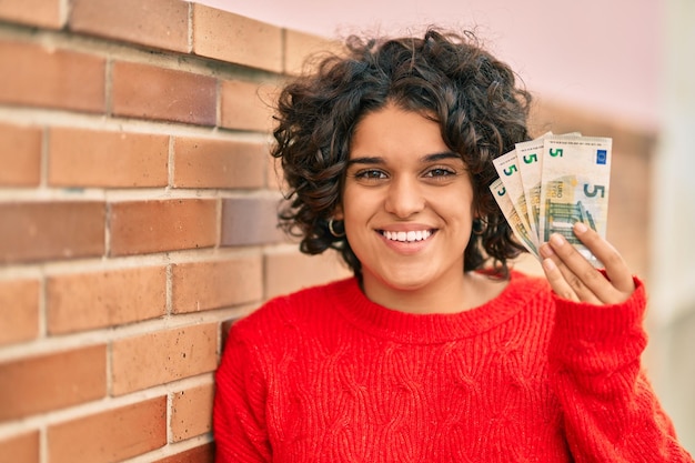 Jovem hispânica sorrindo feliz segurando notas de 5 euros na cidade.