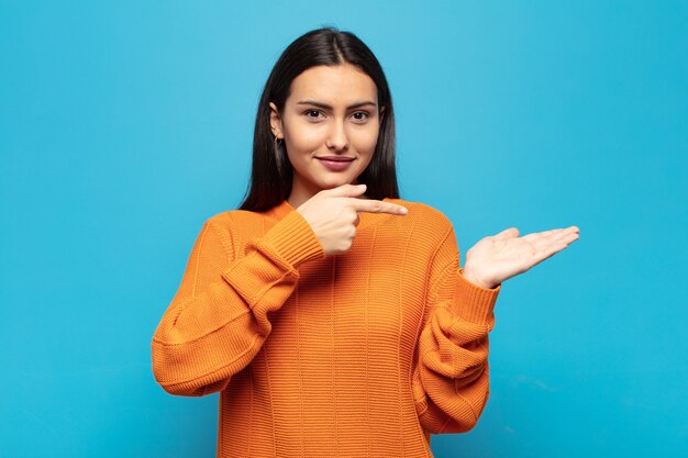 Jovem hispânica sorrindo, sentindo-se feliz, despreocupada e satisfeita, apontando para um conceito ou ideia no espaço da cópia ao lado