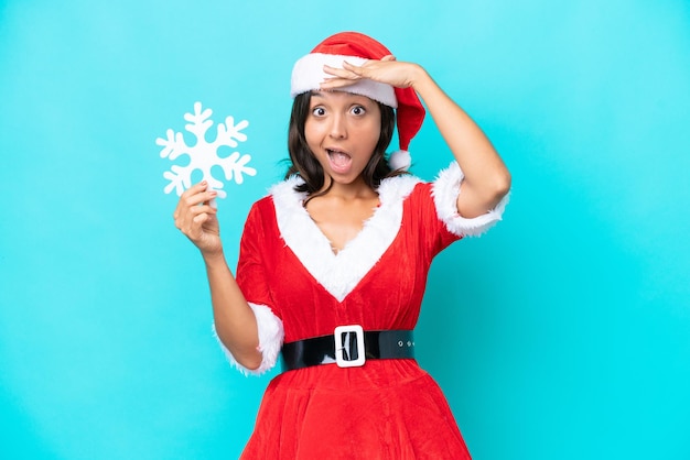 Jovem hispânica vestida de mama noel segurando um floco de neve isolado em fundo azul fazendo gesto de surpresa enquanto olha para o lado