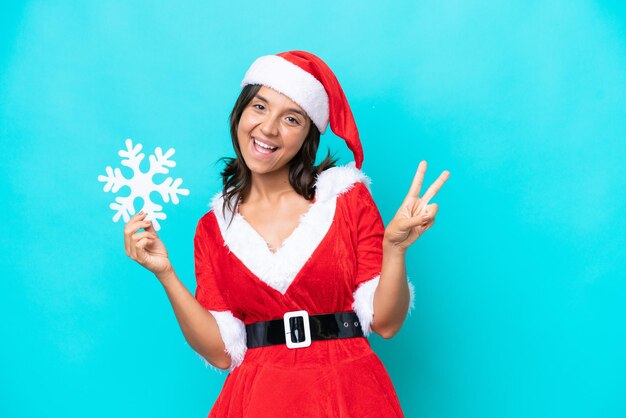 Jovem hispânica vestida de mama noel segurando um floco de neve isolado em fundo azul sorrindo e mostrando sinal de vitória