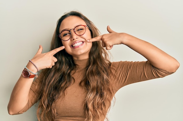 Jovem hispânica vestindo roupas casuais e óculos sorrindo alegre mostrando e apontando com os dedos, dentes e boca conceito de saúde bucal