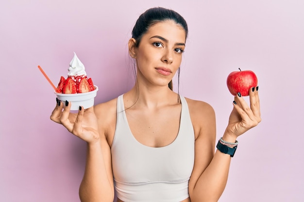 Jovem hispânica vestindo roupas esportivas segurando maçã e sorvete relaxado com expressão séria no rosto simples e natural olhando para a câmera