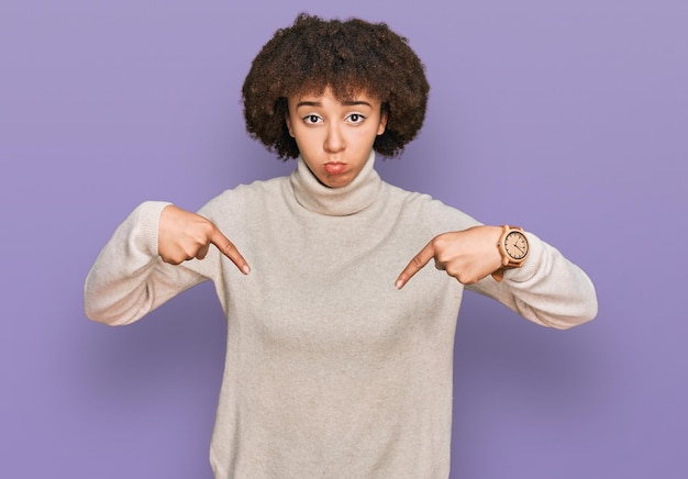 Jovem hispânica vestindo suéter de inverno de lã apontando para baixo parecendo triste e chateada, indicando a direção com os dedos, infeliz e deprimida.