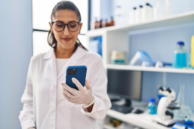 Jovem hispânica vestindo uniforme de cientista usando smartphone no laboratório