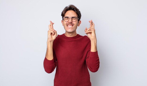 Jovem hispânico bonito sorrindo e cruzando ansiosamente os dois dedos, sentindo-se preocupado e desejando ou esperando boa sorte