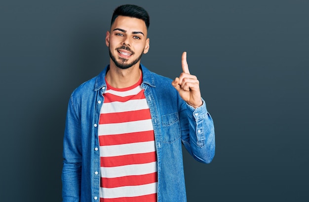 Jovem hispânico com barba vestindo jaqueta jeans casual mostrando e apontando para cima com o dedo número um enquanto sorria confiante e feliz