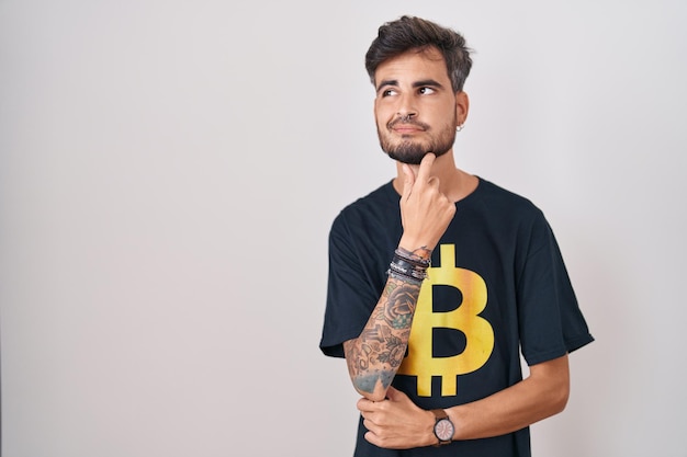 Jovem hispânico com tatuagens vestindo camiseta bitcoin olhando confiante para a câmera sorrindo com braços cruzados e mão levantada no queixo pensando positivo