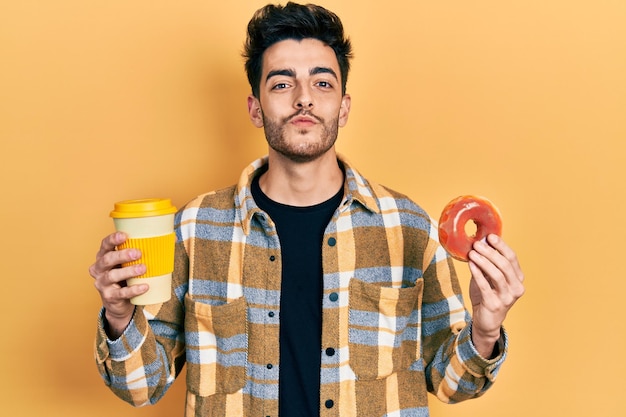 Jovem hispânico comendo rosquinha e bebendo café olhando para a câmera mandando um beijo sendo adorável e sexy. expressão de amor.