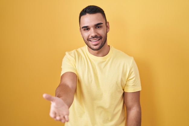 Jovem hispânico de pé sobre fundo amarelo sorrindo alegremente oferecendo palma da mão dando assistência e aceitação