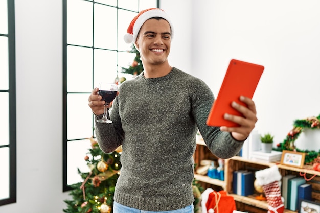 Jovem hispânico faz selfie pelo touchpad bebendo vinho em pé ao lado da árvore de natal em casa