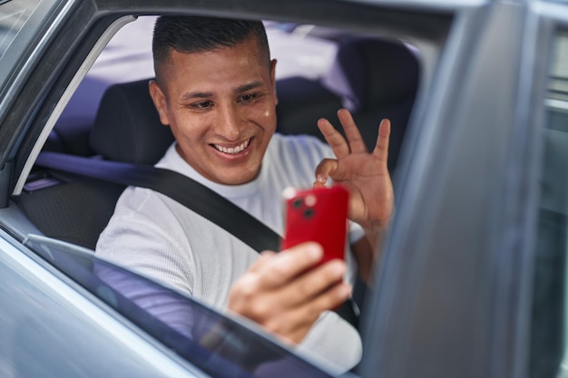 Jovem hispânico fazendo videochamada com smartphone no carro fazendo sinal de ok com os dedos, sorrindo amigável gesticulando excelente símbolo