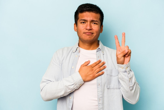 Jovem hispânico isolado em fundo azul fazendo um juramento colocando a mão no peito