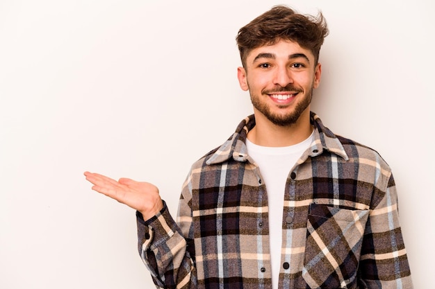 Jovem hispânico isolado no fundo branco, mostrando um espaço de cópia na palma da mão e segurando a outra mão na cintura