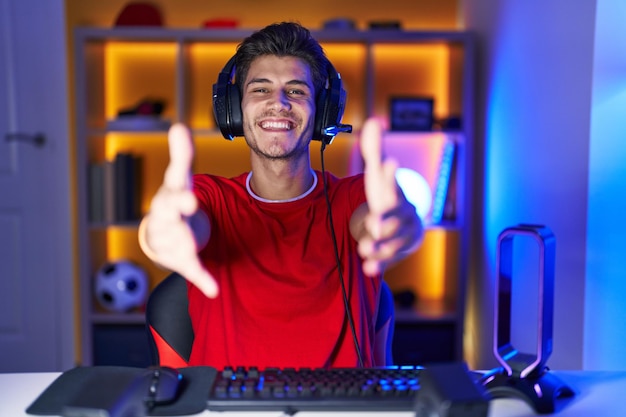 Jovem hispânico jogando videogame olhando para a câmera sorrindo de braços abertos para abraçar a expressão alegre abraçando a felicidade