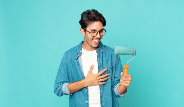 Jovem hispânico rindo alto de uma piada hilária e segurando um rolo de pintura